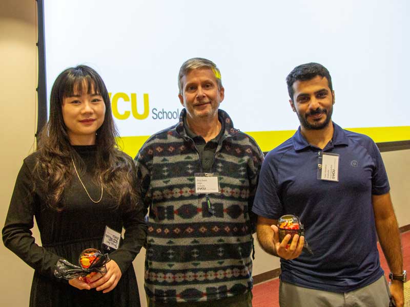 Students Shiya Han and Yasir Alshehry hold awards while standing on either side of past chair Doug Sweet, Ph.D., in front of a lecture hall on Research Day.