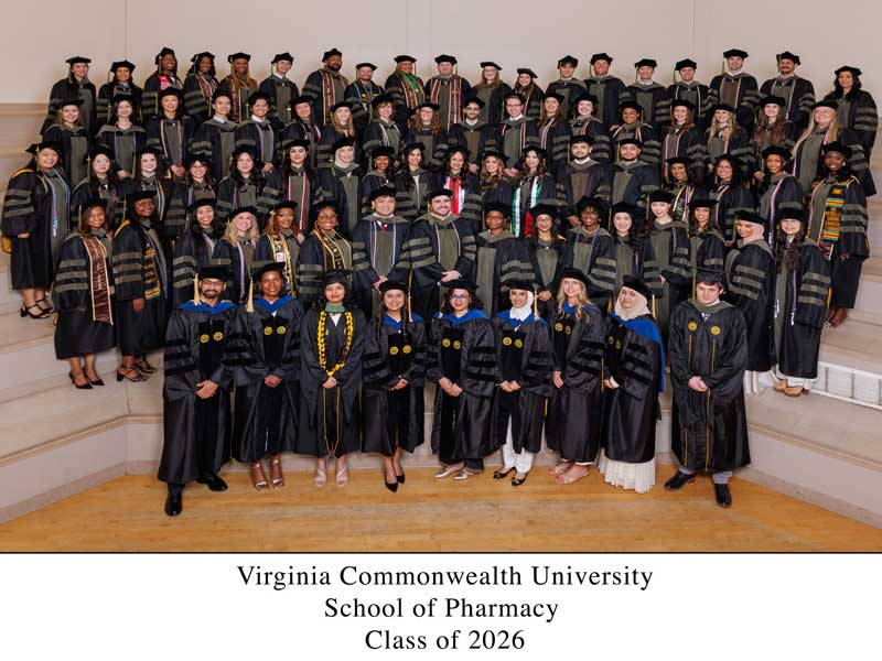 More than 80 members of the VCU School of Pharmacy Class of 2025 in academic regalia (doctoral tams, hoods and gowns) gather on risers for a photo of them celebrating their graduation. Below them, the caption reads 