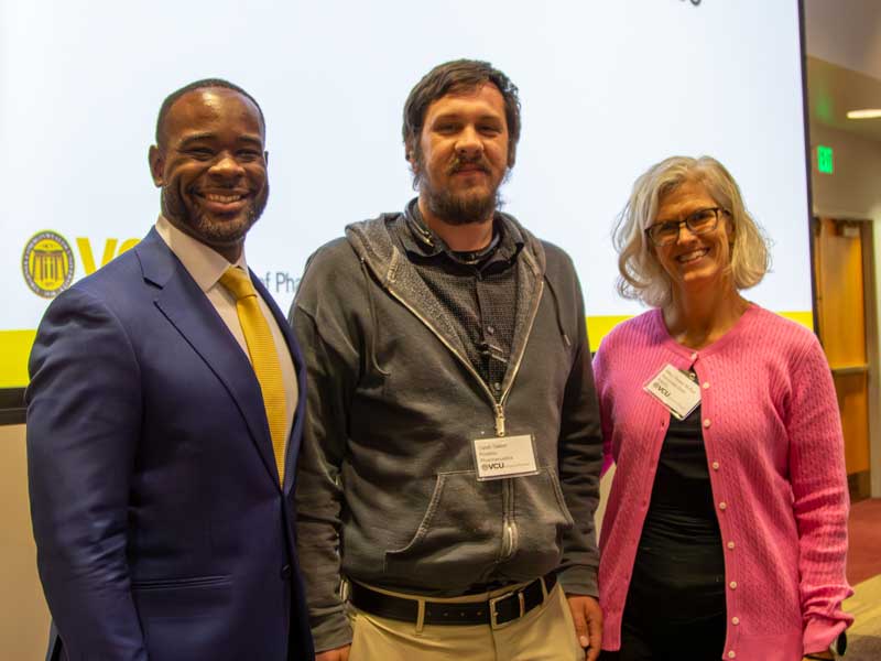 Dean K.C. Ogbonna and Associate Dean Mary Peace McRae stand on either side of postdoc award winner Caleb Dalton, at the front of a lecture hall on Research Day.