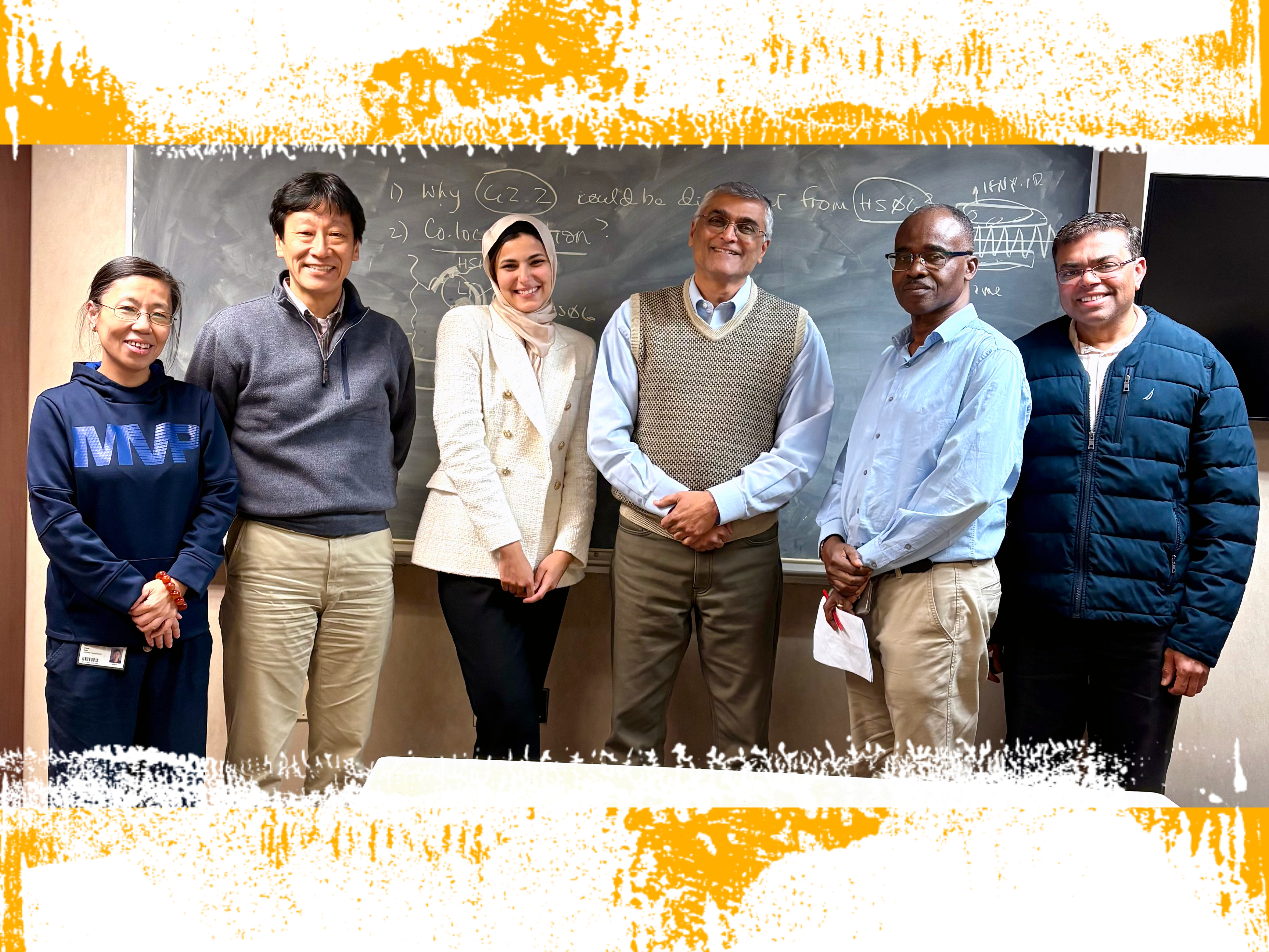 Six people, including a student in formal business attire who has just completed her dissertation defense, smile together for a photo in front of a chalkboard with computational medicinal chemistry formulas written on it.