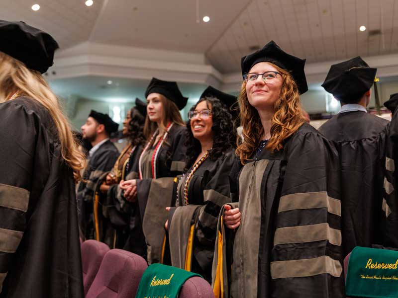 A Class of 2026 graduating student in regalia looks off into the distance with pride and hope ahead of the Hooding and Diploma Ceremony in a large theater-style space.