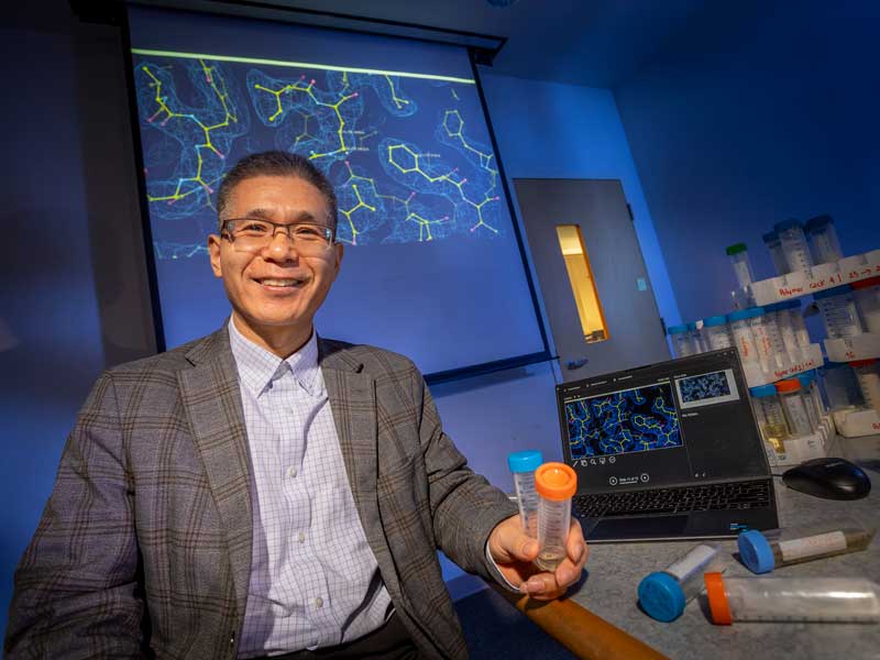 A professor in a dress shirt and blazer sits smiling in his lab in front of a projector with a 2-D representation of the interconnection of molecules while holding two vials of different liquids as additional vials of liquid sit on his desk in front of him