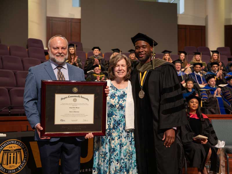 The parents of the late Abby Metzger accept her Pharm.D. diploma from Dean Ogbonna on her behalf on-stage at the VCU School of Pharmacy Hooding and Diploma Ceremony on May 9, 2025.
