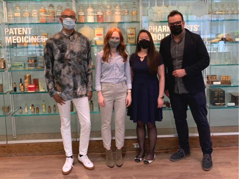 Four masked students stand side by side in front of a wall of pharmacy artifacts during a spring 2021 photo shoot.