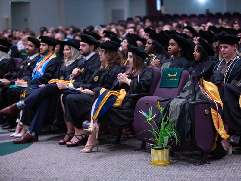A doctoral tam, hood and gown sit next to an empty seat with a reserved sign on it and a potted yellow flower in front of it. The seat was reserved in honor of the late Abby Metzger, a member of the Pharm.D. Class of 2025 who died unexpectedly of natural causes in 2022. The School of Pharmacy and the Class of 2025 chose to honor Abby’s memory with these gifts, and her parents accepted her diploma.