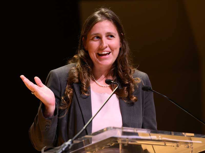 A woman speaks into a microphone at a lectern and gestures with her hand.